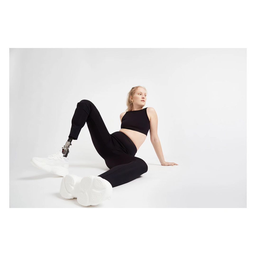 Blonde woman in black athletic wear poses on a white background, showcasing a prosthetic leg and modern white sneakers, looking away.