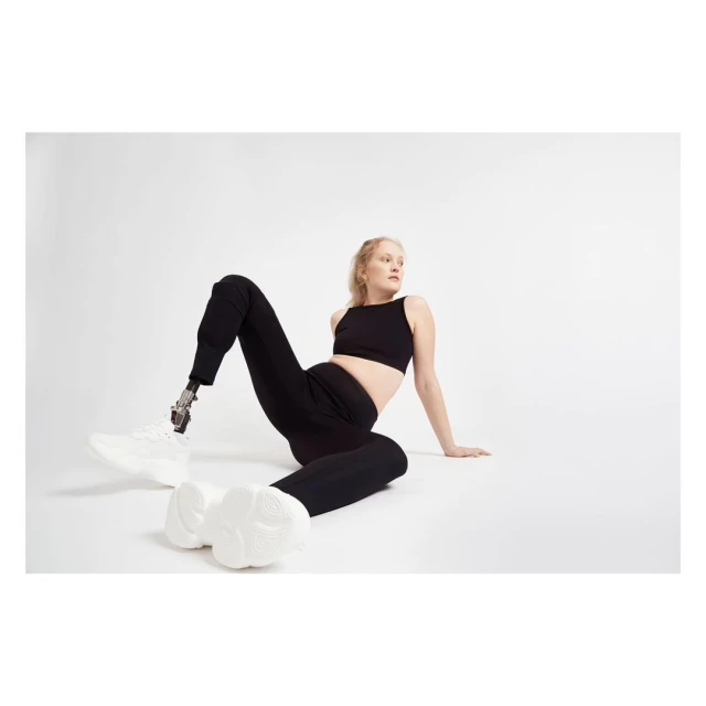 Blonde woman in black athletic wear poses on a white background, showcasing a prosthetic leg and modern white sneakers, looking away.