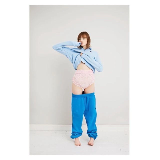 A young woman strikes a pose in a photo studio, wearing blue sweatpants, lace underwear, and a blue sweater, showcasing a blend of casual and chic.