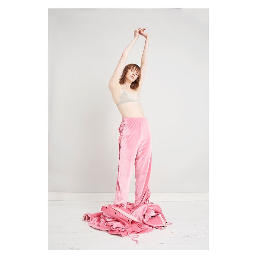 Woman with arms raised, wearing pink velvet pants and a beige bra, standing over a pile of pink clothing in a white room.
