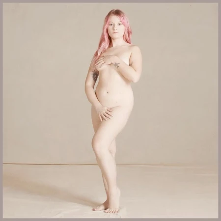Full body studio portrait of a fair-skinned adult female with pink hair, her hands modestly covering her body, against a neutral backdrop.