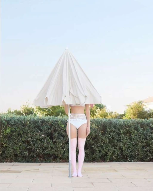 Woman standing outdoors under a white umbrella wearing a white swimsuit and pink thigh-high socks on a sunny day in full shot.