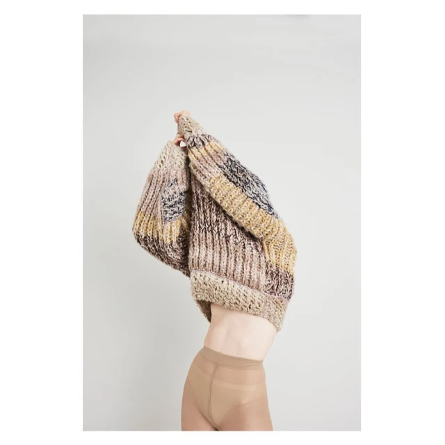 A person partially visible, wearing a multi-colored, knitted sweater, against a light background in a studio setting.