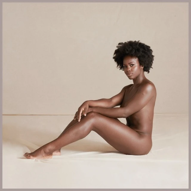 African American woman poses nude on a beige backdrop, seated with knees up and hands resting on legs, with a neutral expression.