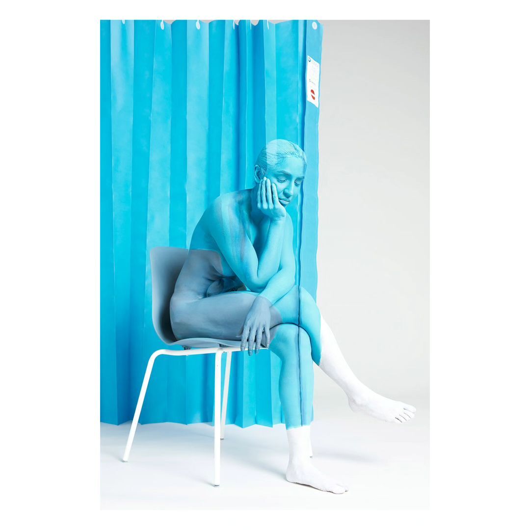 Woman with blue body paint sits on a white chair in front of a blue curtain in a photo studio with white floors and walls.