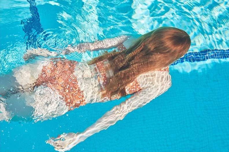 Underwater view of a person swimming in a bright blue pool. The swimmer has long brown hair and a patterned swimsuit in clear water.