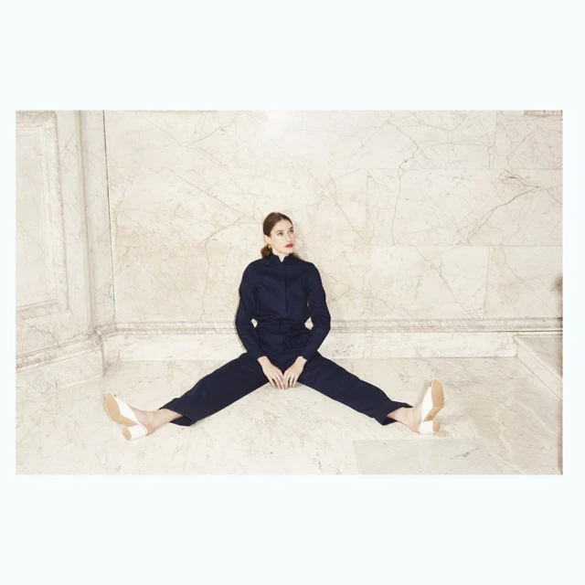 A woman in a dark blue jumpsuit sits with her legs spread against a light marble wall, wearing light-colored heels in a studio shot.