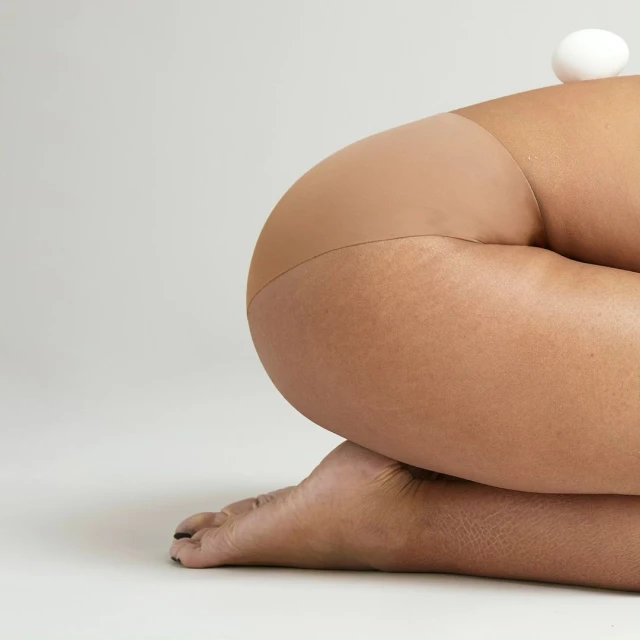 Close-up of a person's lower body kneeling, wearing tan tights, with an egg resting on their back against a light gray background.