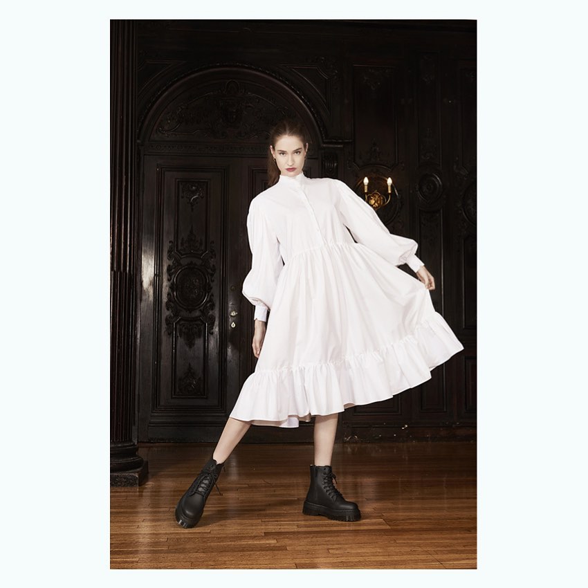 A young adult woman in a white dress and black boots stands indoors in front of a dark wood door and column, fashion shot.