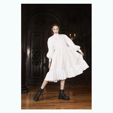 A young adult woman in a white dress and black boots stands indoors in front of a dark wood door and column, fashion shot.