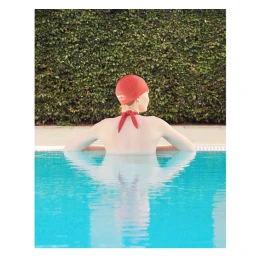 Woman wearing a red swim cap in a turquoise outdoor swimming pool with a tall green hedge backdrop
