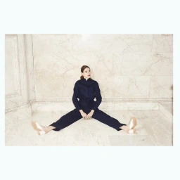 Model wearing dark jumpsuit and white mules sitting on a marble floor against an ornate wall