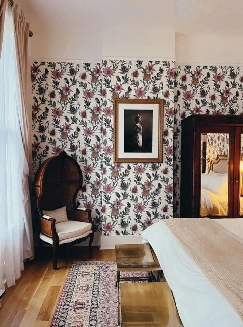 Bedroom with floral wallpaper, an ornate chair, a framed picture, and a bed with a tan throw, creating a cozy and elegant atmosphere.