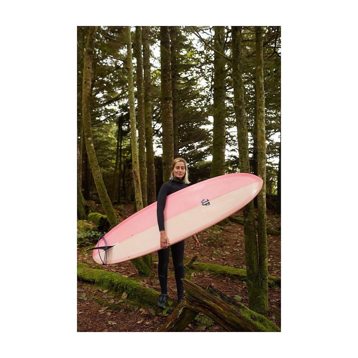 A woman in a wetsuit stands in a forest, holding a pink and white surfboard, with tall trees and mossy ground creating a natural backdrop.