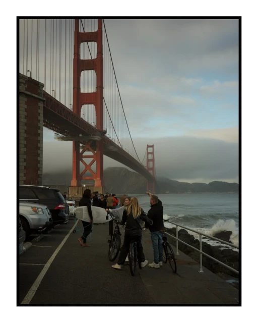 A group of people with bicycles and a surfboard standing in front of the Golden Gate Bridge, with cars parked nearby.
