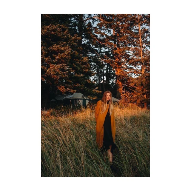 A lone woman in a yellow coat strolls through a sunlit meadow in front of a stand of trees, evoking a tranquil, natural atmosphere.