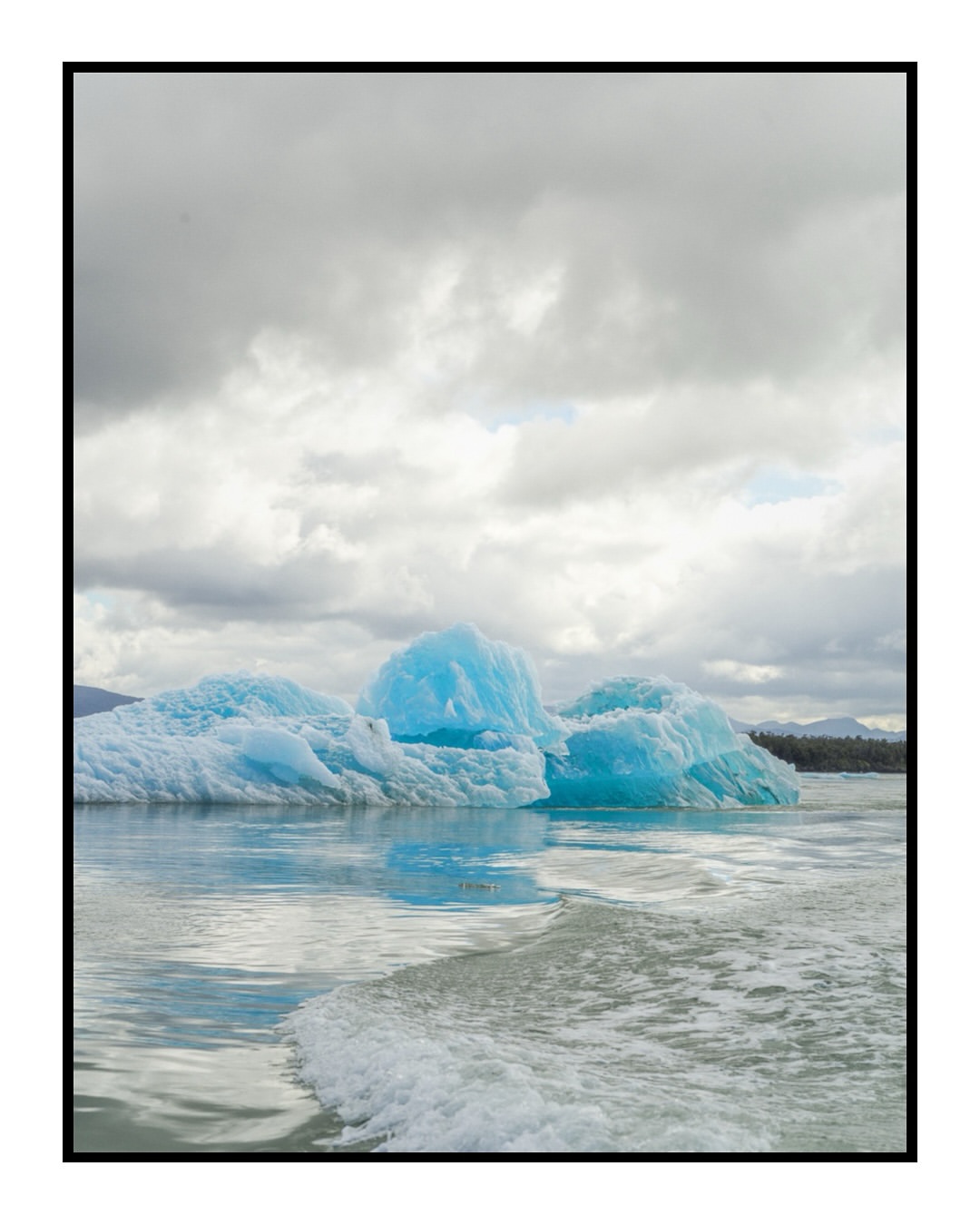 Icy blue icebergs float in calm waters under a cloudy sky, with distant dark land and gentle waves visible.