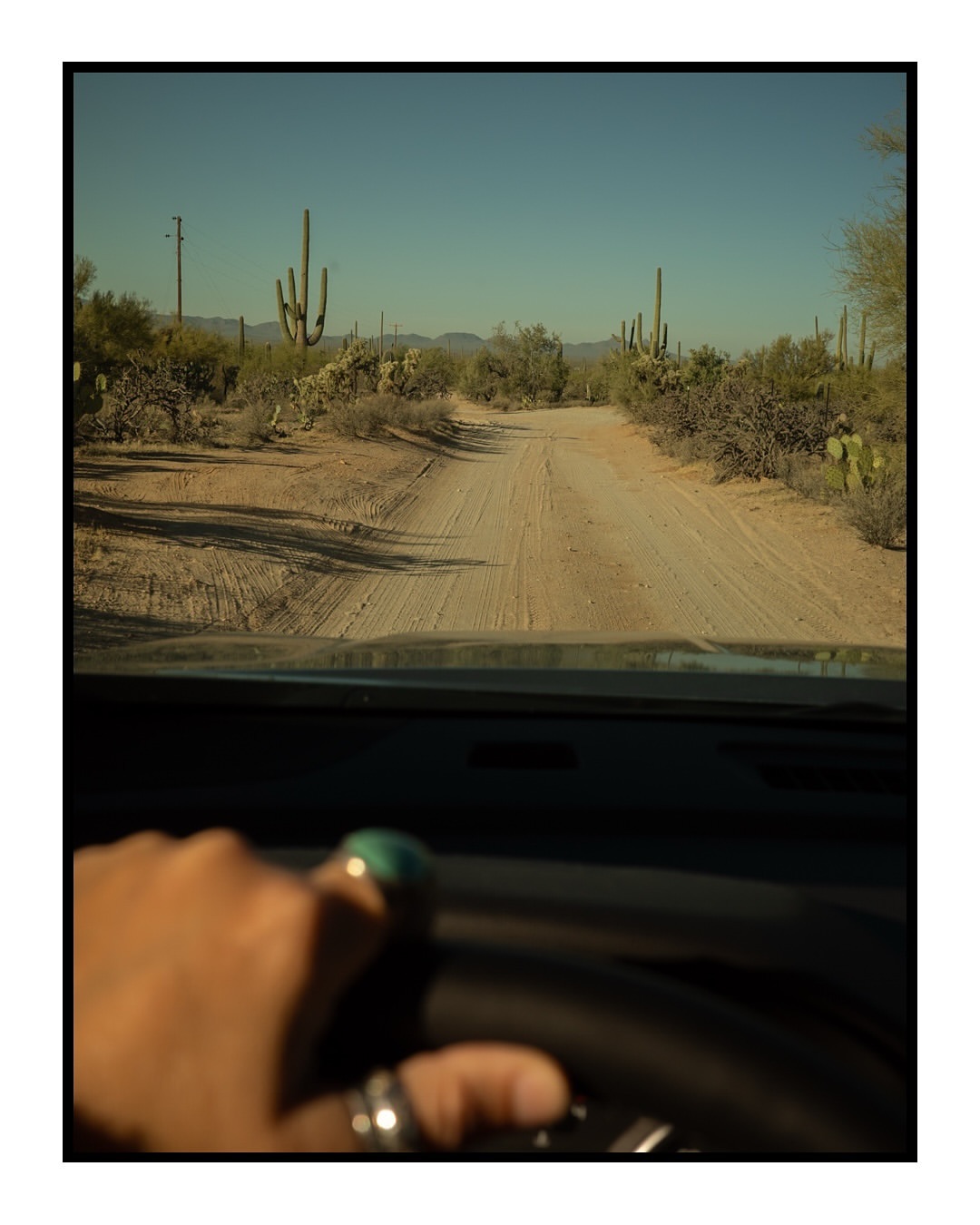 Driving through the Arizona desert with saguaro cacti under a clear blue sky, steering wheel and a hand with rings visible.
