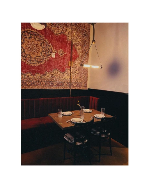 Interior view of restaurant with wooden table set with glasses and plates, flanked by red velvet booth and chairs, with patterned wall rug.