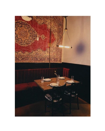 Interior view of restaurant with wooden table set with glasses and plates, flanked by red velvet booth and chairs, with patterned wall rug.