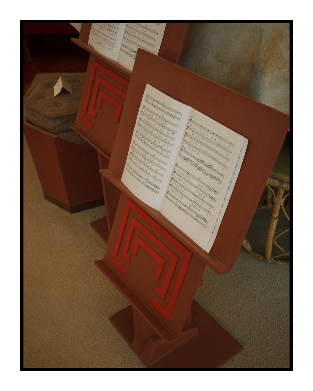 Two vintage music stands with sheet music are displayed indoors on a carpeted floor. The stands feature a geometric design and warm colors.