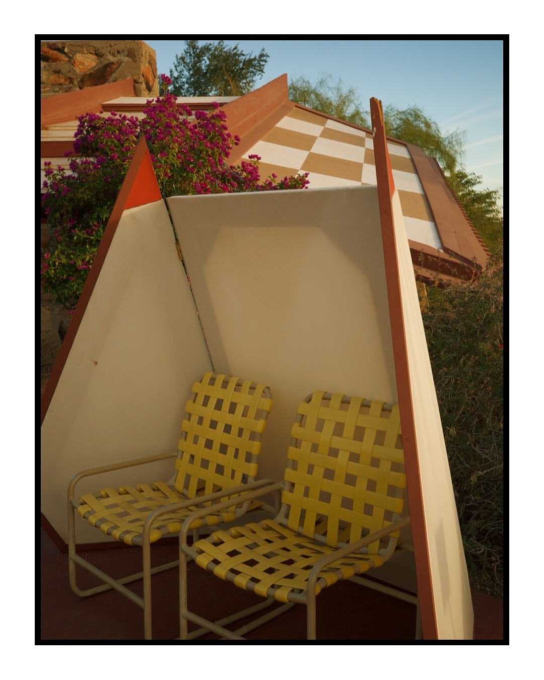 Two yellow wicker chairs sit beneath an angular, cream-colored awning in a sunny outdoor setting. Decorative flora and geometric architectural details.