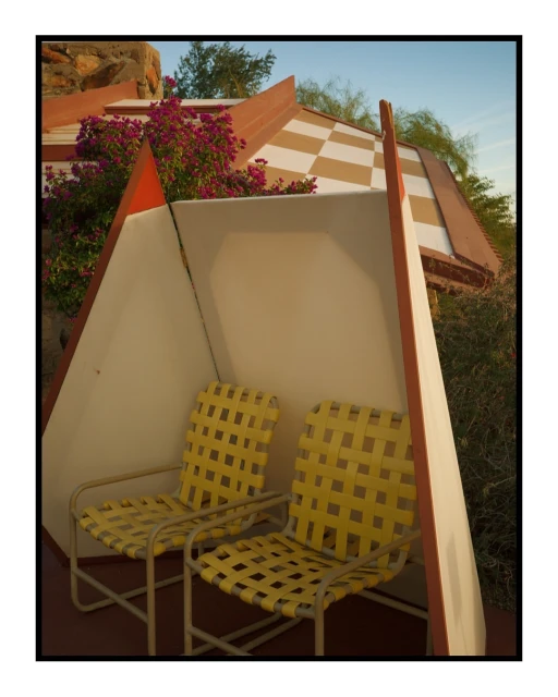 Two yellow wicker chairs sit beneath an angular, cream-colored awning in a sunny outdoor setting. Decorative flora and geometric architectural details.
