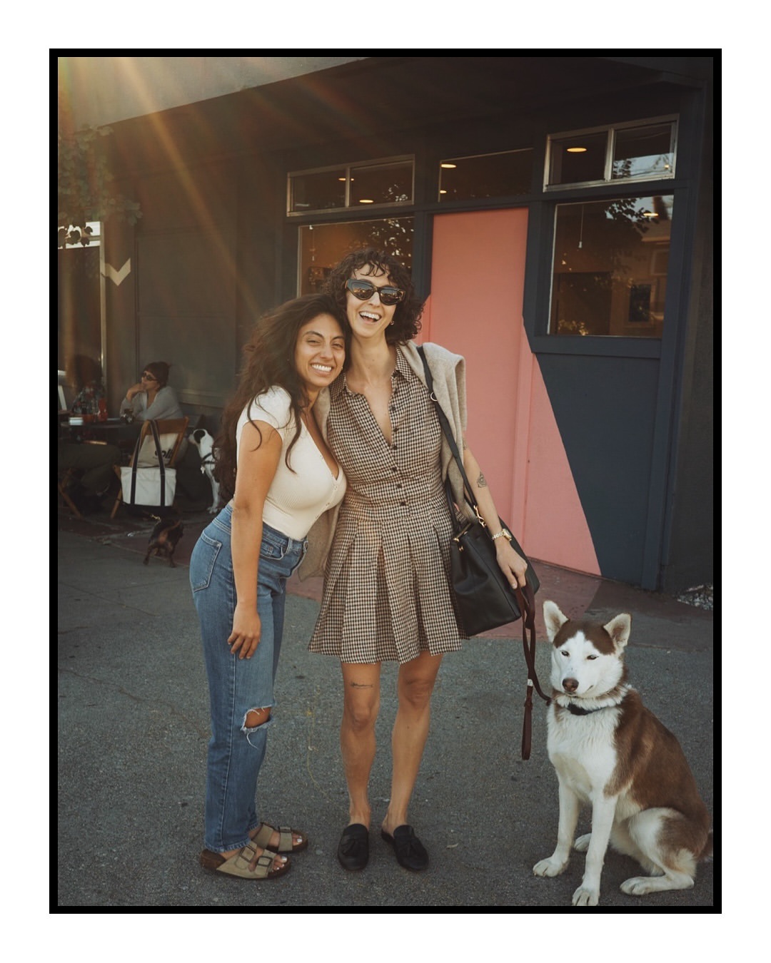 Two women and a dog pose outside a building with a pink door on a sunny day. One woman wears jeans, the other a plaid dress.