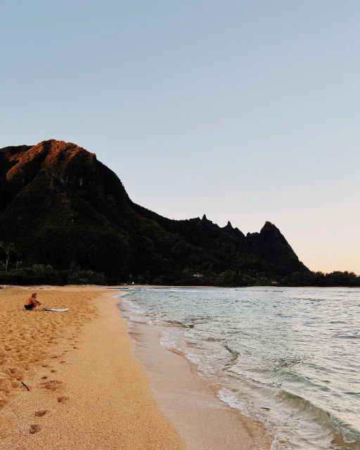 Seascape view of a person sitting on a sandy beach next to a surfboard, facing the ocean, with a large mountain in the background.