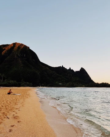 Seascape view of a person sitting on a sandy beach next to a surfboard, facing the ocean, with a large mountain in the background.