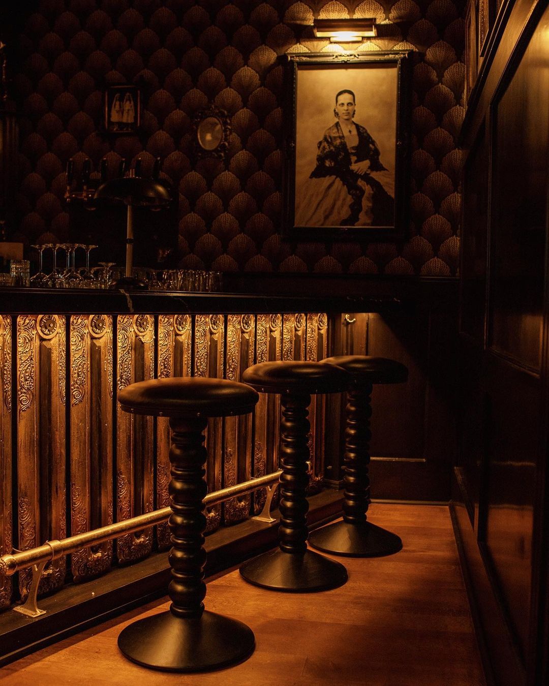 Interior view of a dimly lit bar area with three dark-stained wooden stools in the foreground, illuminated by warm overhead lighting.