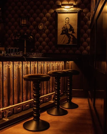 Interior view of a dimly lit bar area with three dark-stained wooden stools in the foreground, illuminated by warm overhead lighting.