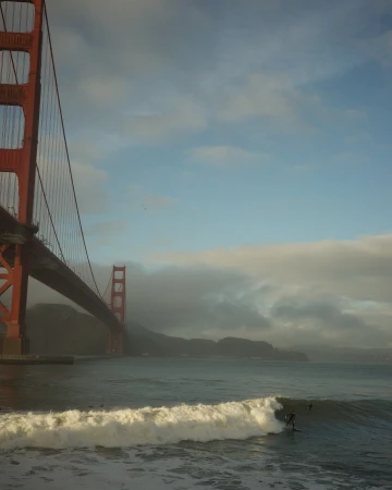 Golden Gate Bridge with surfers riding waves in the foreground on a foggy day, creating a classic San Francisco scene.