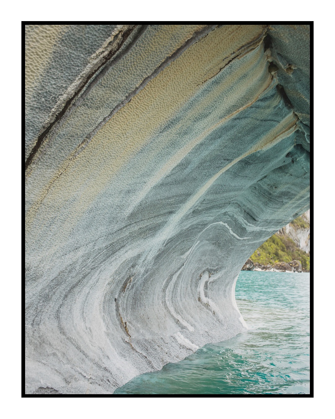 Striking geological formation featuring a cave with marble-like rock textures, set against turquoise water and distant greenery.