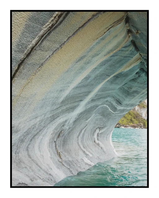 Striking geological formation featuring a cave with marble-like rock textures, set against turquoise water and distant greenery.
