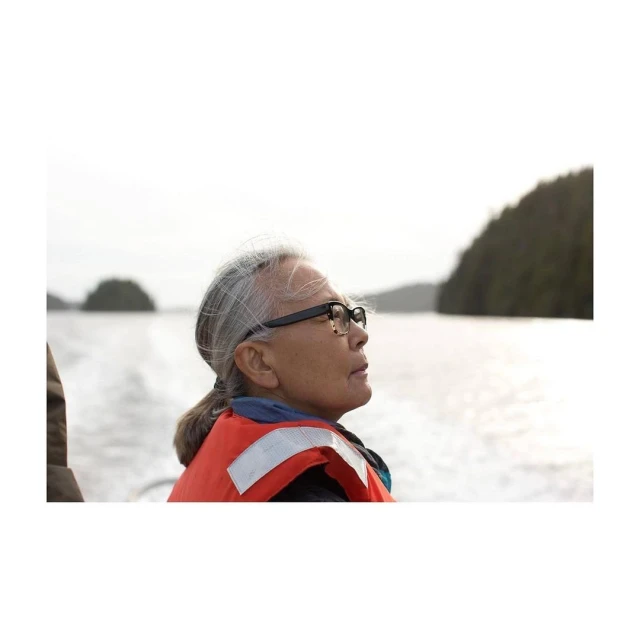 Profile of senior woman with grey hair and glasses wearing a life vest on a boat, looking towards the forested coastline.