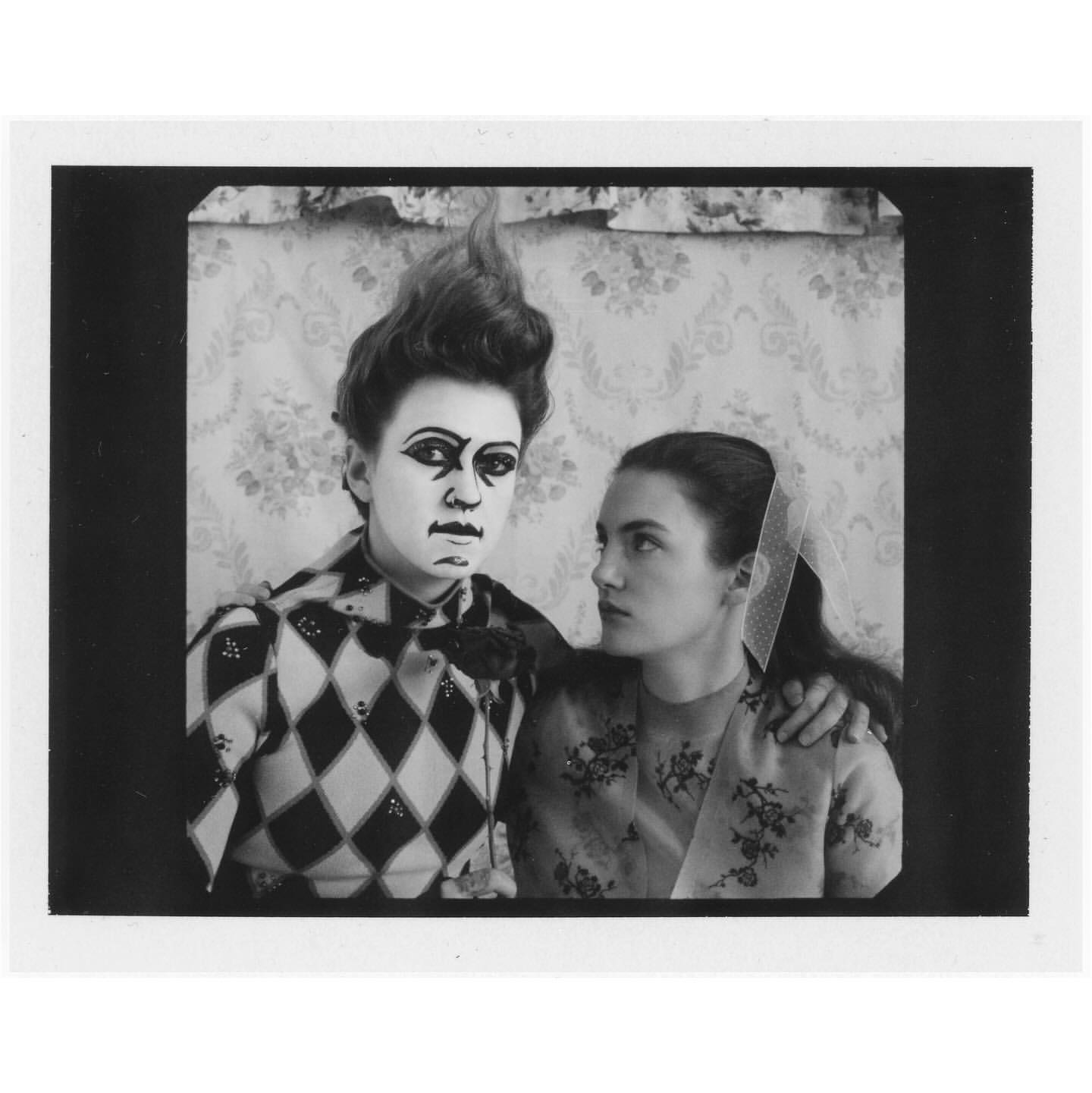 Two women pose indoors: One with clown makeup and harlequin patterned top, the other in floral top and netting veil, vintage black and white photograph.