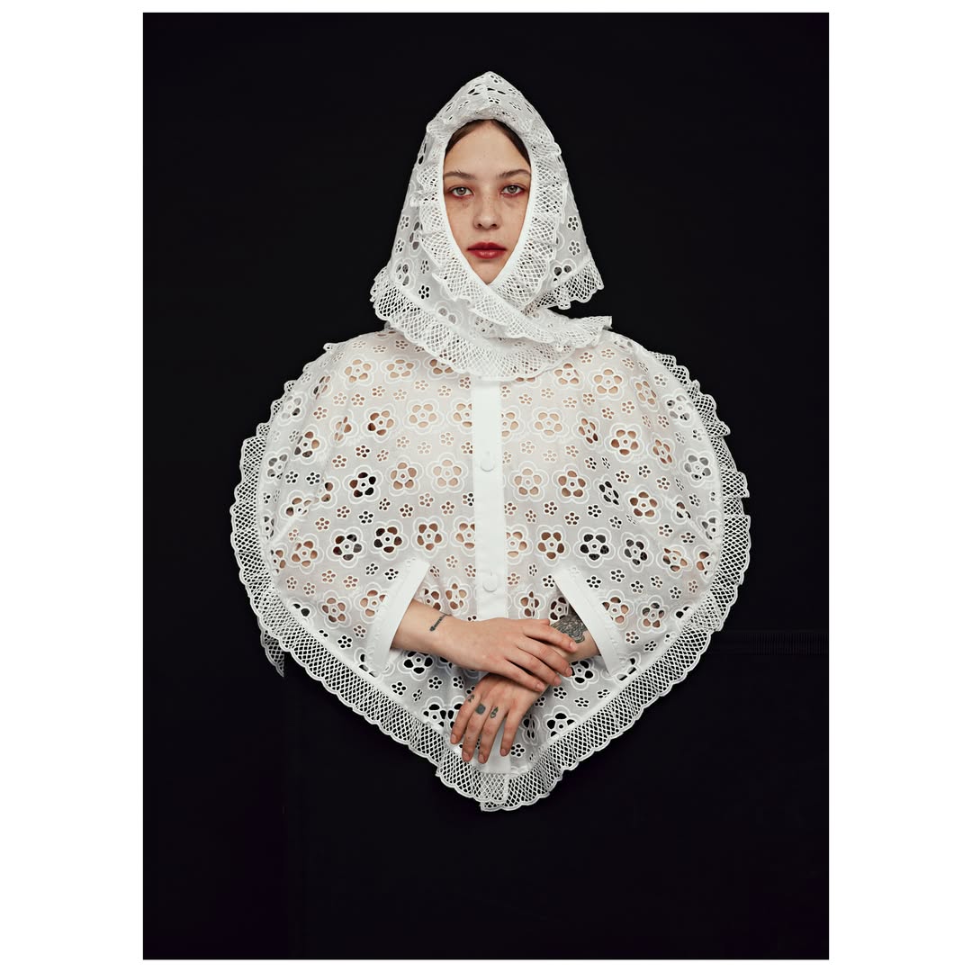 Studio shot of a fair-skinned person wearing a white, eyelet lace bonnet and cape, against a dark background. Fashion, beauty, and studio lighting.