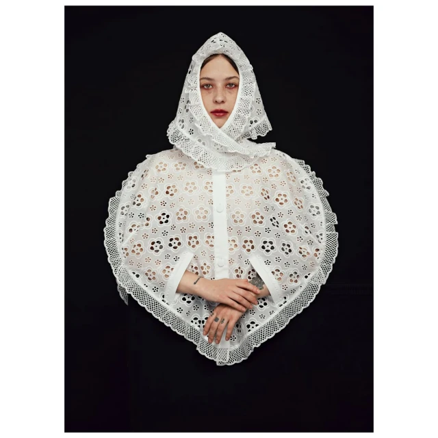 Studio shot of a fair-skinned person wearing a white, eyelet lace bonnet and cape, against a dark background. Fashion, beauty, and studio lighting.