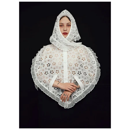 Studio shot of a fair-skinned person wearing a white, eyelet lace bonnet and cape, against a dark background. Fashion, beauty, and studio lighting.