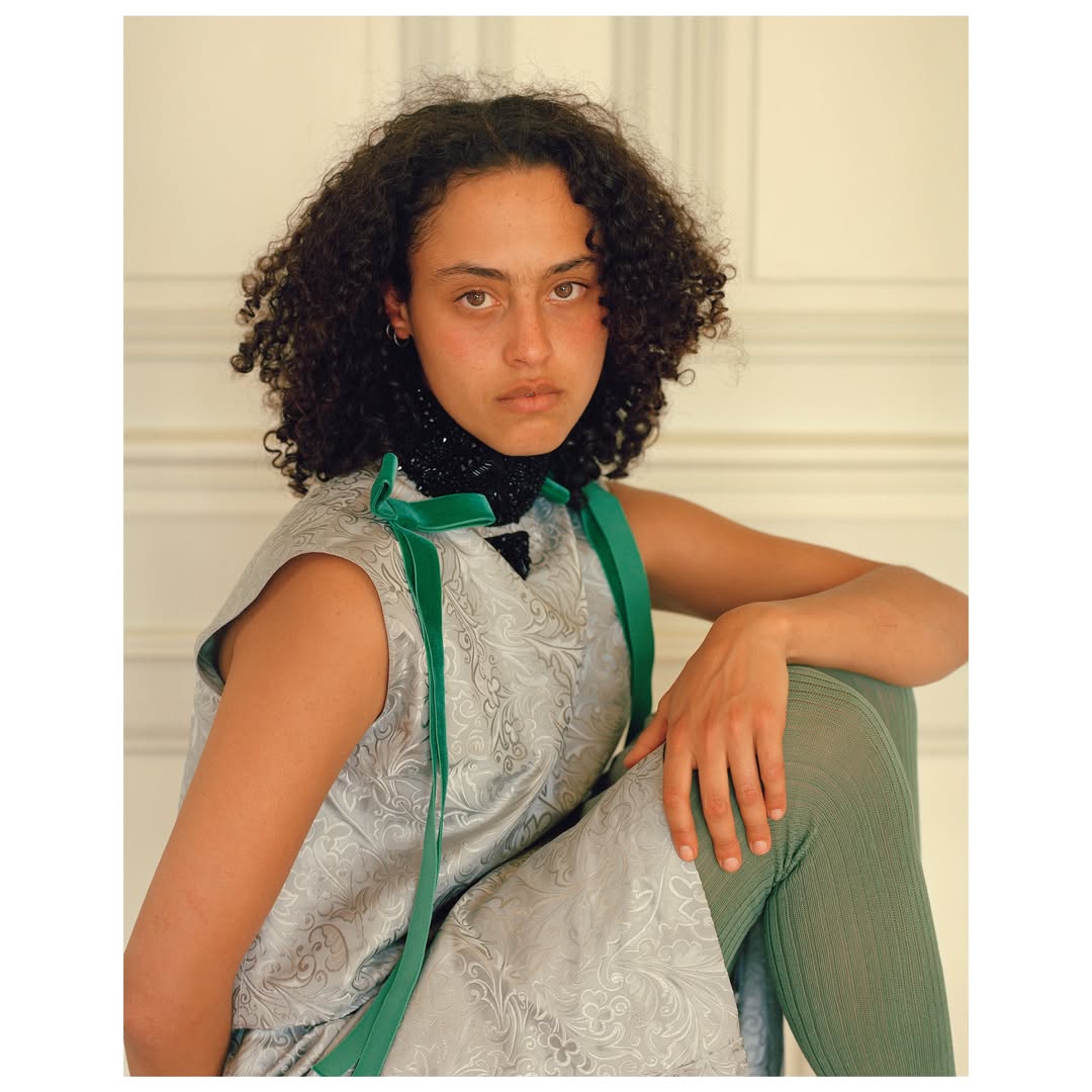 Young woman with curly hair poses indoors wearing a sleeveless silver and green dress, tights, and a beaded collar, looking at the camera.