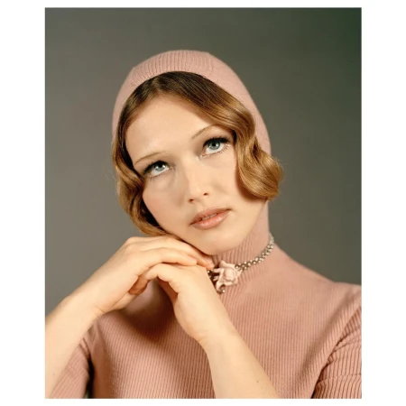 Portrait of a woman in a pink turtleneck and knit cap, looking up with clasped hands and silver floral necklace against a grey backdrop.