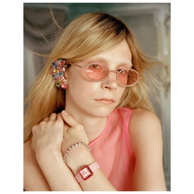 A stylish young woman wearing round pink-tinted glasses, adorned with colorful jeweled earrings and bracelet, poses in a close-up studio portrait.