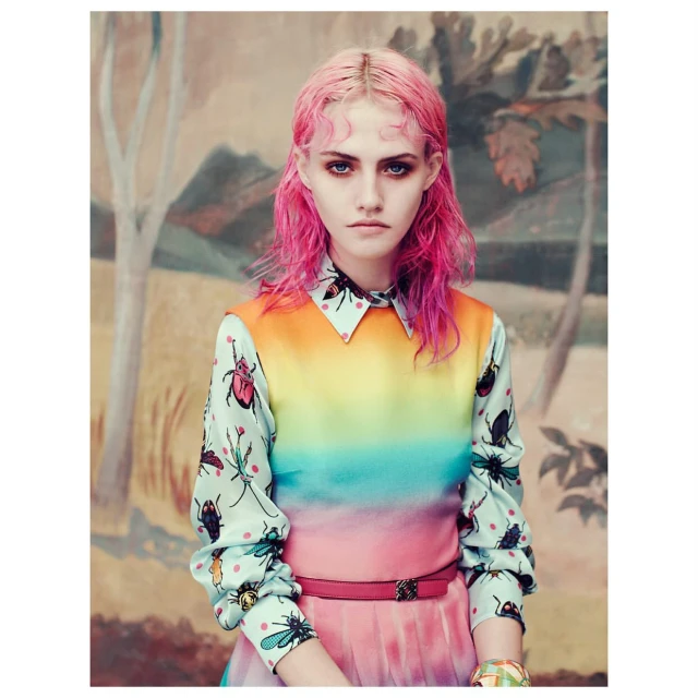 Portrait of a young adult female fashion model with pink hair wearing a colorful insect-themed blouse in front of a painted backdrop.