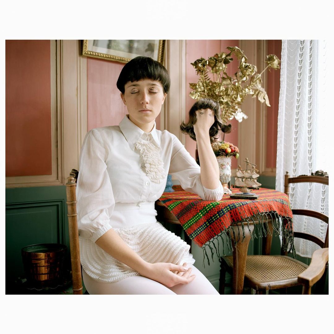 Indoor portrait of a young woman with closed eyes in a white ruffled shirt, seated at a table with a colorful tablecloth and gold decor.