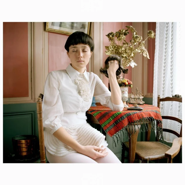 Indoor portrait of a young woman with closed eyes in a white ruffled shirt, seated at a table with a colorful tablecloth and gold decor.