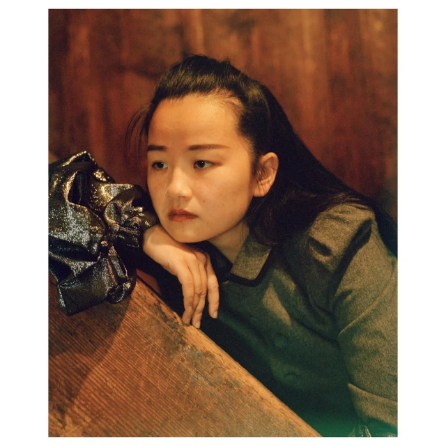 Portrait of an Asian woman in a gray jacket with long hair, resting her face on her hand near a shiny handbag on a wooden surface.