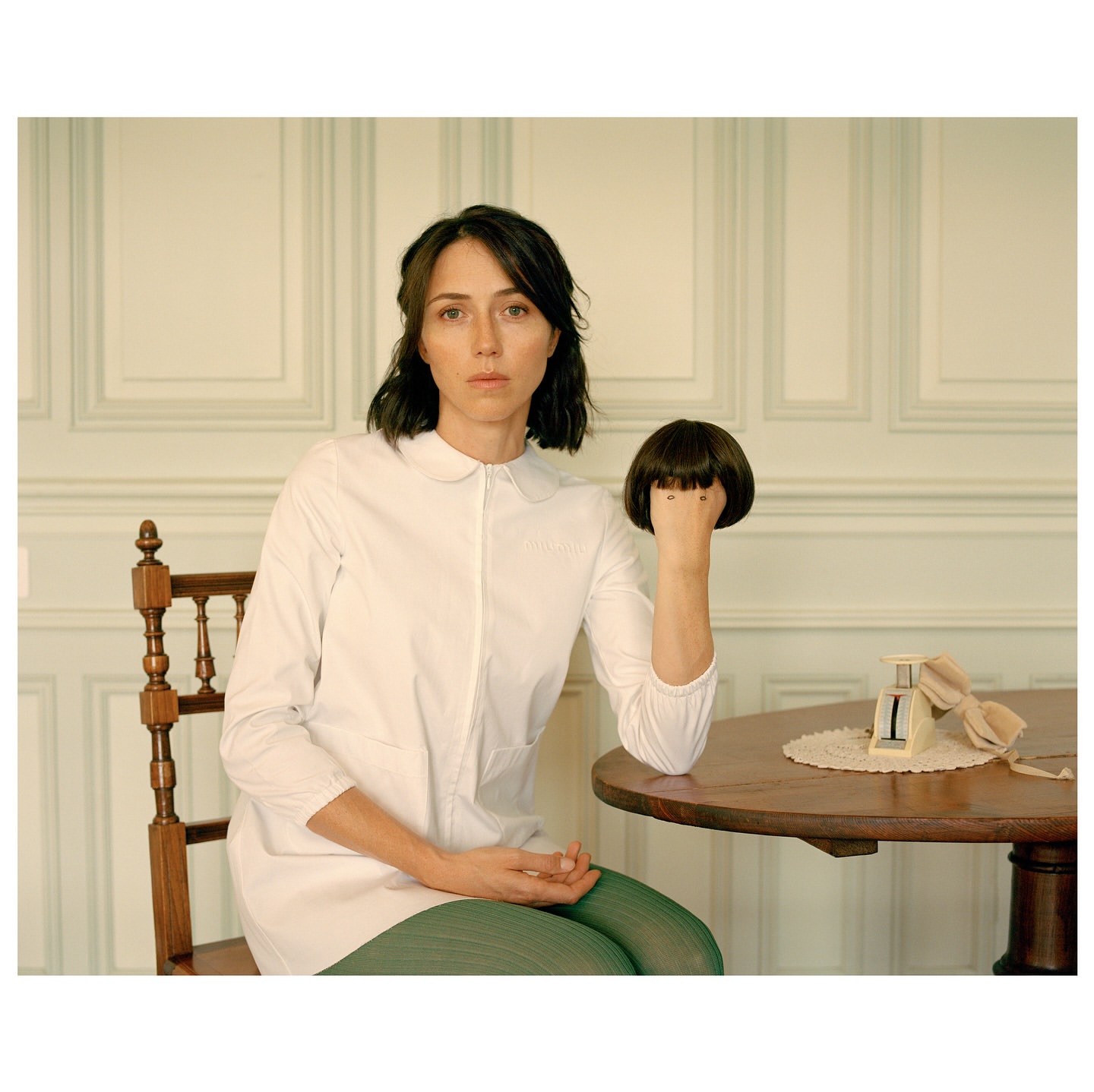 Indoor portrait of a woman seated at a table, holding a dark brown wig. The table also has a calendar and lace doily.
