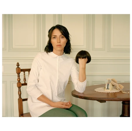Indoor portrait of a woman seated at a table, holding a dark brown wig. The table also has a calendar and lace doily.