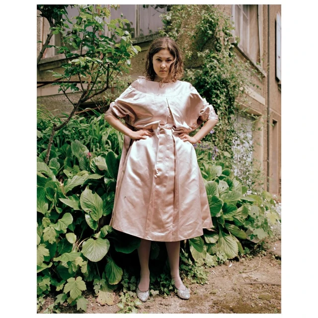 Young woman in a pink couture dress stands confidently outdoors near lush green plants and a stone building with ivy, fashion.